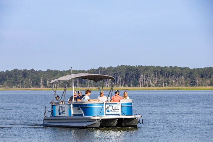 A group of people on a blue pontoon boat on a lake with a forested shoreline in the background.