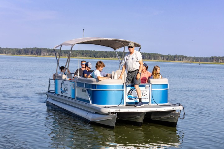 People on a blue pontoon boat on a calm lake with a forested shoreline.